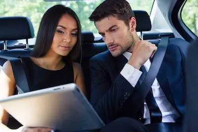 Two people in formal attire sit in a car, looking intently at a tablet.