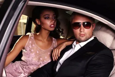 A woman in a pink dress and a man in a black suit with sunglasses sit in the backseat of a car.