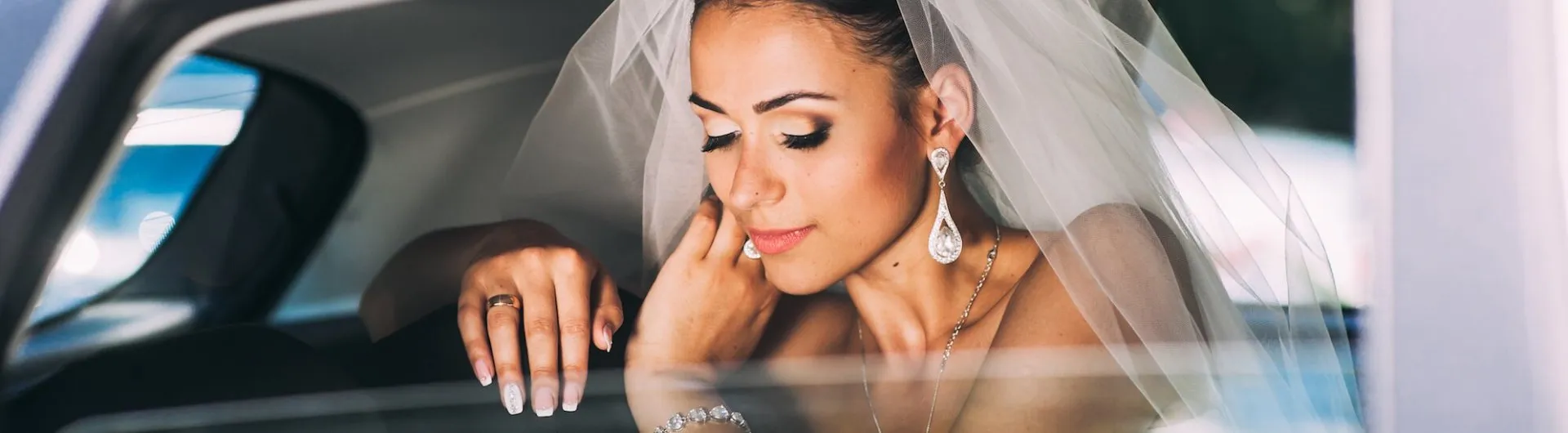 Bride in a wedding dress and veil sits in a car, touching her face gently, with eyes closed and an expression of calm.