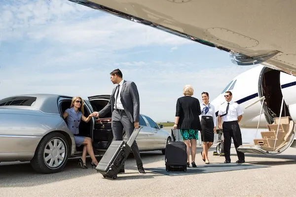 A woman exits a car as a man and two others stand near a private jet, one holding luggage. Two crew members are positioned close to the jet's stairs.