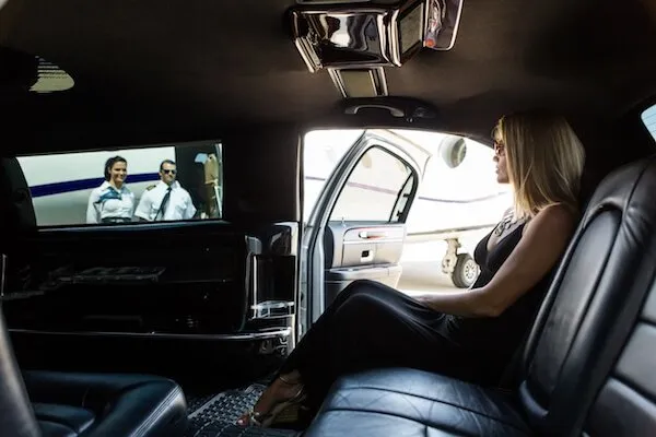Person in a black dress sits in a car, watching a movie on a screen. An airplane is visible outside through the open door.