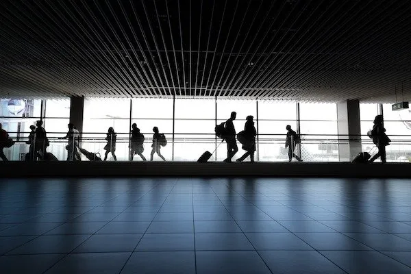 Silhouetted travelers with luggage walk through an airport terminal.