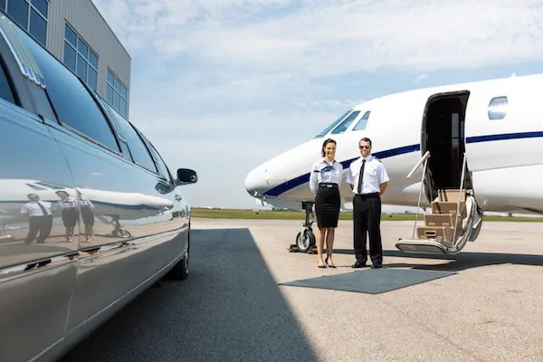 Two pilots stand beside a private jet on an airport tarmac, with a limousine parked nearby under a clear sky.