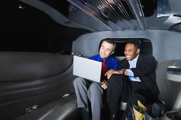 Two men in business attire are sitting in a limousine, looking at a laptop and smiling.