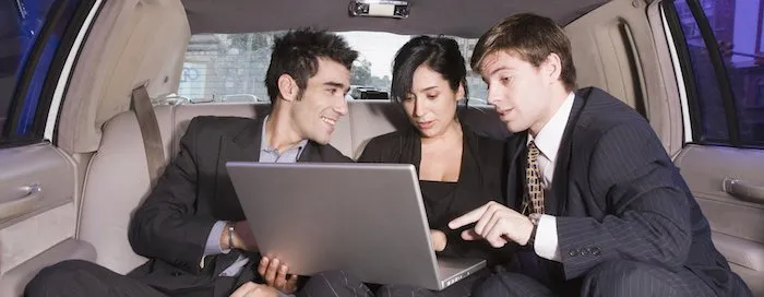 Three people in business attire sit in the backseat of a car, looking at a laptop.