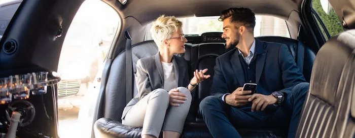 Two people in formal attire sit in the backseat of a car, talking. One holds a smartphone.