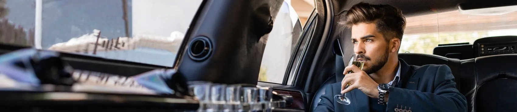 A man in a suit sits in a car, holding a glass and gazing out the window.