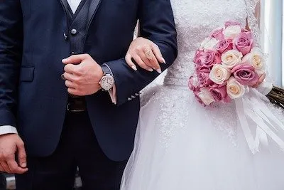 A bride and groom stand arm in arm; the bride holds a bouquet of pink and white roses.