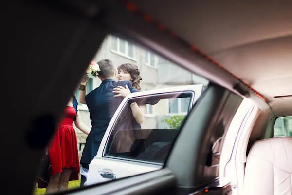 View from inside a car showing a person in a formal blue outfit embracing a person in a blue dress near an open door. Another person in a red dress stands nearby.